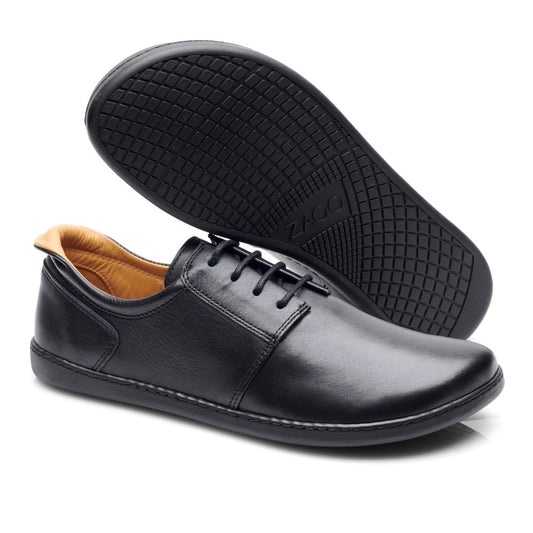 Ein Paar PIQUANT Nappa Black Schnürschuhe aus schwarzem Leder mit sichtbaren Nähten und hellbrauner Innenseite, von der Seite und von unten sichtbar. Die Sohle weist ein geometrisches Griffmuster mit eingeprägtem ZAQQ-Branding auf.