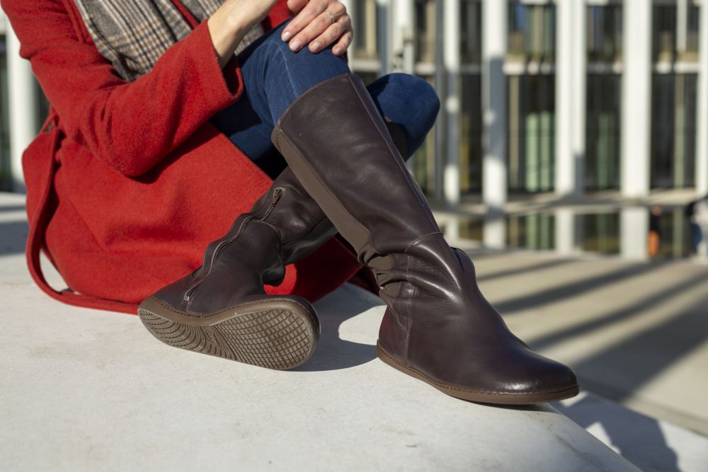 Eine Person sitzt auf einer Betonfläche und trägt einen roten Mantel, blaue Jeans und ZAQQs QATCH Nappa Brown hohe Damenstiefel. Sonnenlicht wirft Schatten auf den Boden, während im Hintergrund ein modernes Gebäude steht.