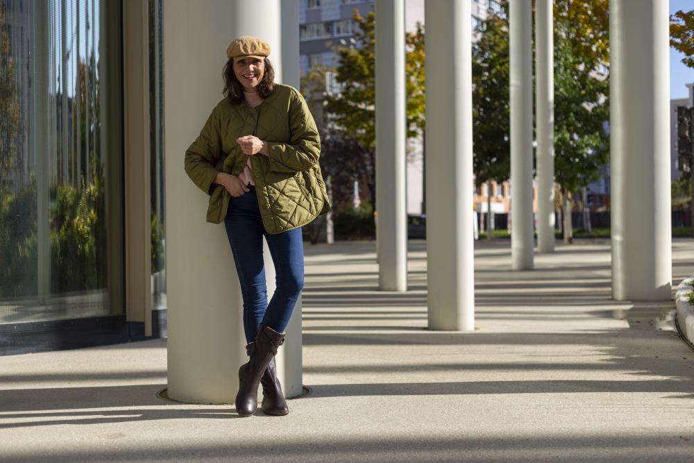 Eine Person in einer Steppjacke ATTRAQT Winter Brown von ZAQQ, Jeans und Winterstiefeln lehnt an einer Säule in einer städtischen Umgebung. Sie trägt eine beigefarbene Mütze und lächelt, genießt den Tragekomfort, während Bäume und moderne Gebäude die Szene einrahmen.