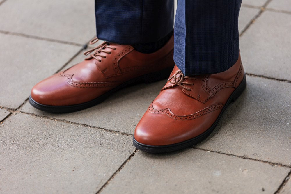 Eine Person trägt die polierten, cognacfarbenen BRIQ Brogue-Schuhe von ZAQQ mit ihren aufwendigen Details, kombiniert sie mit einer marineblauen Hose und steht selbstbewusst auf einem gepflasterten Gehweg.