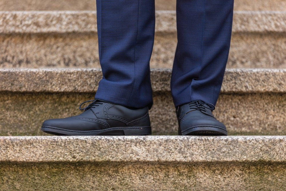 Eine Person ist auf einer Steintreppe zu sehen, die dunkelblaue Hosen und BRIQ Brogue Vegan Black Abendschuhe von ZAQQ trägt. Diese Schuhe, die für ihr klassisches Design und ihre dekorativen Nähte bekannt sind, sind mit einer rutschhemmenden Sohle ausgestattet. Das Bild konzentriert sich auf die Unterschenkel und Füße der Person.