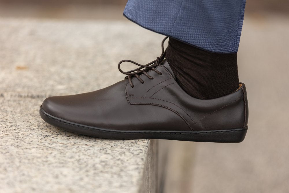 Nahaufnahme einer Person, die ZAQQs PEAQ dunkelbraunen Halbschuh mit Schnürung aus Nappaleder trägt, kombiniert mit schwarzen Socken und blauen Hosen. Der Fuß steht auf einer Betonoberfläche, was das Business-Casual-Design und die Textur der Schuhe hervorhebt.