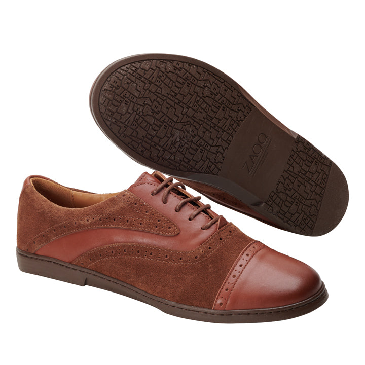 Ein Paar QENTLE Cognac-Schuhe der Marke ZAQQ im Oxford-Stil aus braunem Leder und Wildleder mit Brogue-Details. Ein Schuh steht aufrecht und zeigt sein aufwendiges Oberdesign, während der andere angewinkelt ist und seine elegant gemusterte Sohle zeigt, was eine Handwerkskunst zeigt, die an Nappaleder-Handwerk erinnert.