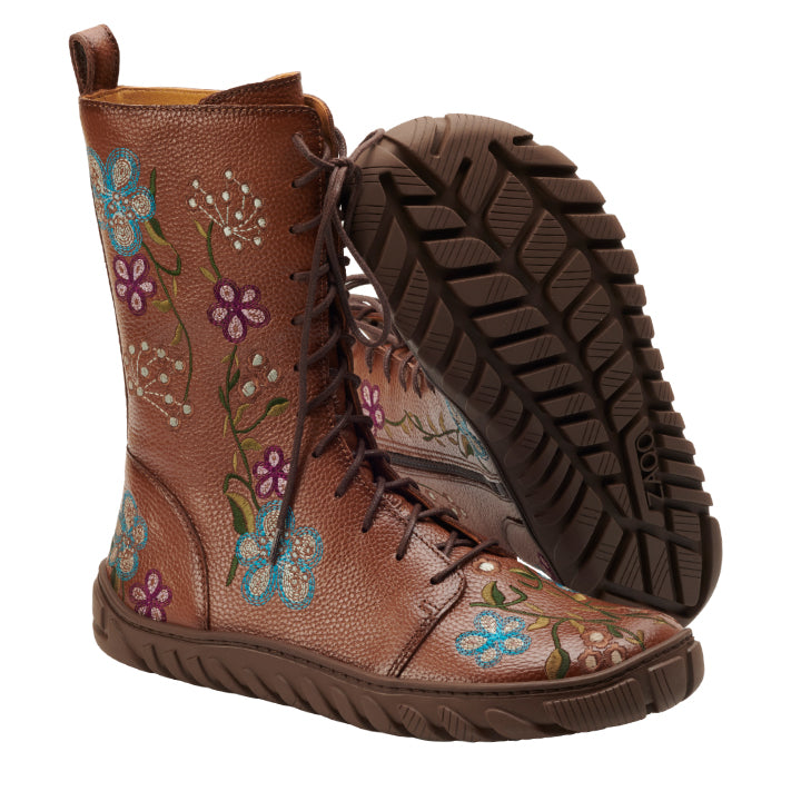 Die DOQS Flower-Kollektion von ZAQQ umfasst braune Schnürstiefel, die mit aufwendigen Blumenstickereien verziert und mit robusten Sohlen ausgestattet sind. Ein Stiefel steht aufrecht, während der andere angewinkelt ist, um sein Profil hervorzuheben und so eine Eleganz mit Retro-Charme einzufangen.