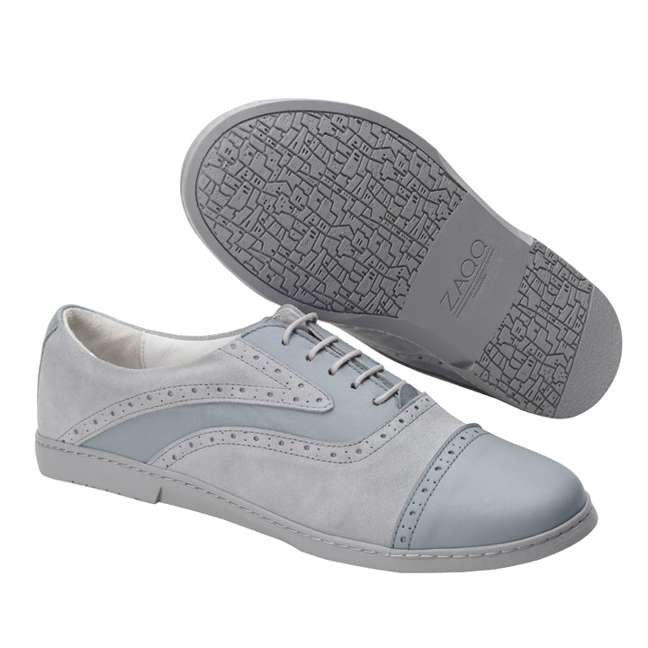 Wir präsentieren den QENTLE Grey von ZAQQ: ein Paar stilvoller Business-Schuhe, fachmännisch gefertigt aus einer Kombination aus Velours und Nappaleder in Hellgrau und Blau. Diese Schuhe zeichnen sich durch ein perforiertes Design mit einer kontrastierenden Zehenkappe aus. Ein Schuh ist elegant flach gelegt, um die Draufsicht hervorzuheben, während der andere seine Sohle mit einem komplizierten geometrischen Profilmuster zur Schau stellt.