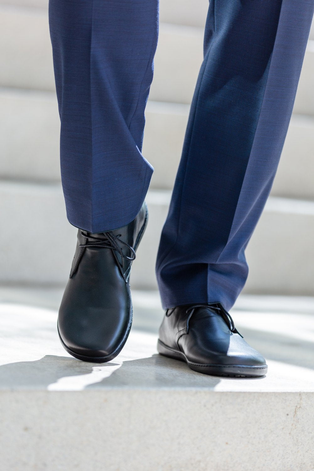 Eine Person in dunkelblauen Hosen und eleganten schwarzen ARQ-Schuhen von ZAQQ steigt eine helle Betontreppe hinab. Ihre anmutigen Schritte werden durch den eleganten Kontrast der polierten schwarzen Schuhe vor dem hellen Hintergrund betont. Das Bild konzentriert sich auf ihre Unterschenkel und Füße und vermittelt einen Eindruck von Vornehmheit.