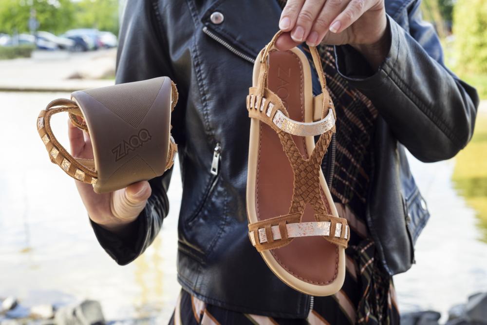 Eine Person in einer Lederjacke hält ein Paar BLINQ Brown-Sandalen und betont damit ihre Flexibilität. Eine Sandale ist hochgekrempelt, während die andere ihr feminines Design mit silberfarbenen Riemen zur Schau stellt. Das ZAQQ-Logo ist auf der Sohle vor einer verschwommenen Außenkulisse sichtbar.