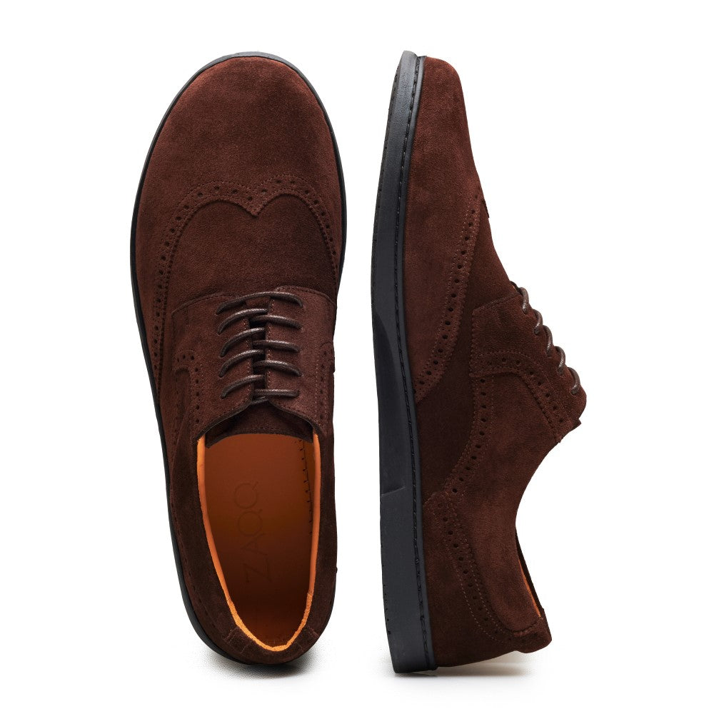 Ein Paar „BRIQ Brogue Velours Brown“ von ZAQQ wird ausgestellt. Ein Schuh ist von oben zu sehen, während der andere von der Seite präsentiert wird. Sie sind aus französischem Kalbleder gefertigt, verfügen über detaillierte Nähte und ein Schnürdesign, wobei eine orangefarbene Innensohle hervorschaut.