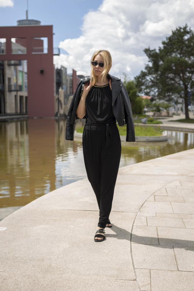 Eine Frau mit schwarzer Sonnenbrille und einem eleganten ZAQQ BROOQE Black-Outfit, mit einer lässig über die Schultern gelegten Jacke, schlendert selbstbewusst einen geschwungenen Weg neben einem Gewässer entlang. Ihre femininen Sandalen verleihen Eleganz, während sich moderne Gebäude und Bäume gegen den teilweise bewölkten Himmel abzeichnen.