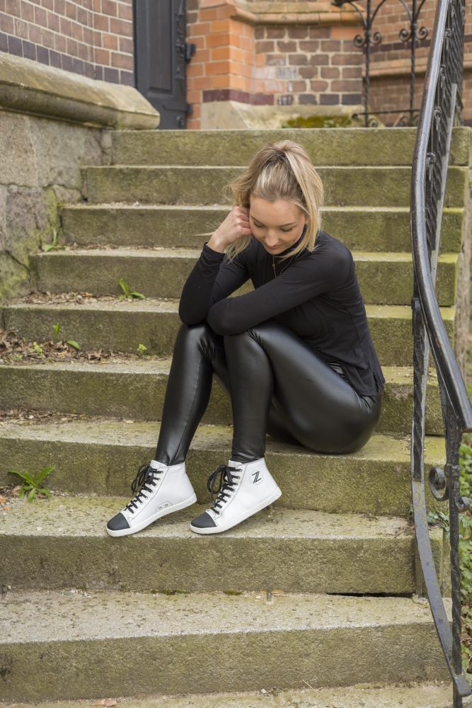 Eine Frau mit blondem Haar sitzt auf einer Außentreppe und stützt sich auf eine Hand. Sie trägt ein schwarzes Oberteil, eine schwarze Lederhose und weiße CHUQQS Sneakers aus Nappaleder der Marke ZAQQ. Die Treppe ist von einem schmiedeeisernen Geländer gesäumt, im Hintergrund ist eine Backsteinmauer zu sehen.