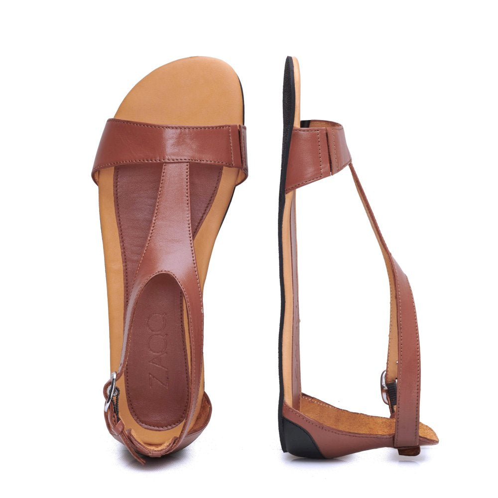 Ein Paar CLIQ Cognac-Sandalen von ZAQQ, hergestellt aus hochwertigem Nappaleder in einem satten Braunton, verfügt über ein stilvolles T-Riemen-Design mit flacher Sohle. Eine Sandale wird flach hingelegt, um die Draufsicht zu zeigen, und die andere steht aufrecht, um ihr schickes Riemchensandale-Seitenprofil zu zeigen. Diese Sandalen verkörpern Schlichtheit und Eleganz.