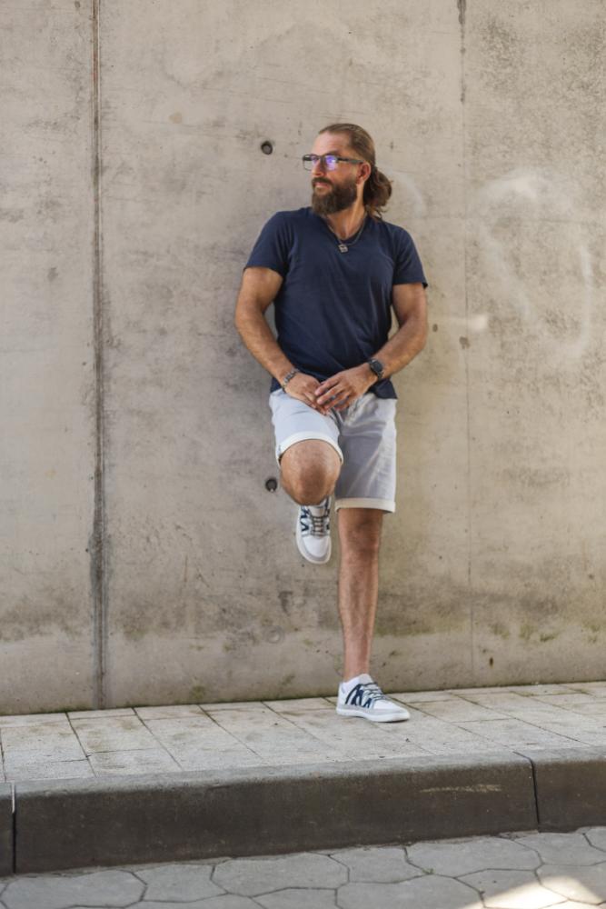 Ein bärtiger Mann mit Sonnenbrille lehnt an einer Betonwand. Er trägt ein marineblaues Hemd, helle Shorts und DRAQE White Grey Sneakers von ZAQQ. Er steht auf einem gefliesten Bürgersteig und hat ein Bein angewinkelt, was eine lässige, entspannte Pose ergibt.