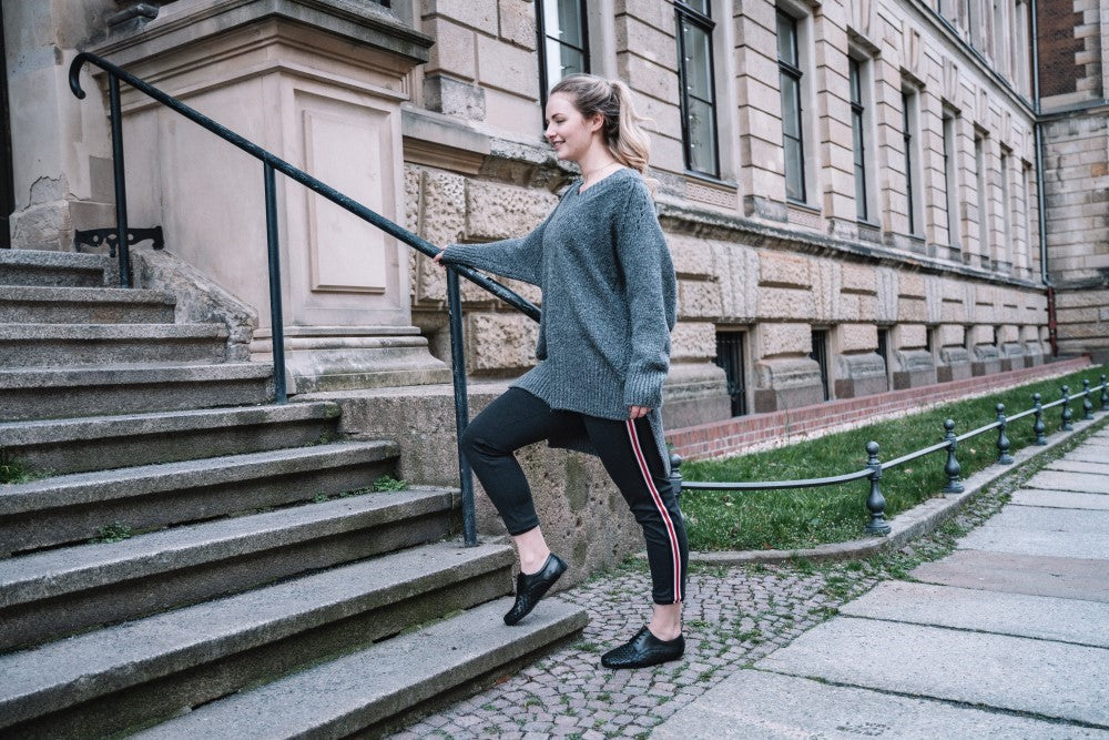 Eine Person in einem grauen Pullover und dem IQON Plait Black von ZAQQ trainiert auf einer Außentreppe neben einem Altbau mit großen Fenstern. Mit zeitloser Eleganz lächelt die Person beim Aufsteigen und genießt die Bewegungsfreiheit, während sie sich am Geländer festhält.