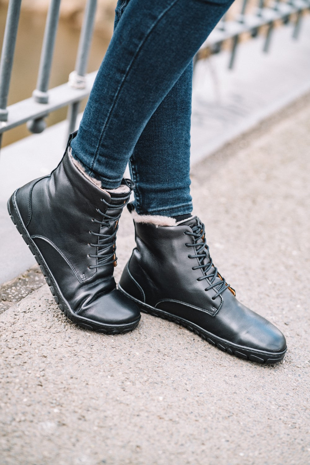 Eine Person, die QUINTIC Winter Black-Stiefel von ZAQQ und Blue Jeans trägt, steht auf einem Bürgersteig neben einem Metallzaun. Die Schnürsenkel der Stiefel sind ordentlich gebunden und die Jeans ist an den Knöcheln leicht hochgekrempelt, wodurch das detaillierte Design der QUINTIC Winter Black-Stiefel sichtbar wird.