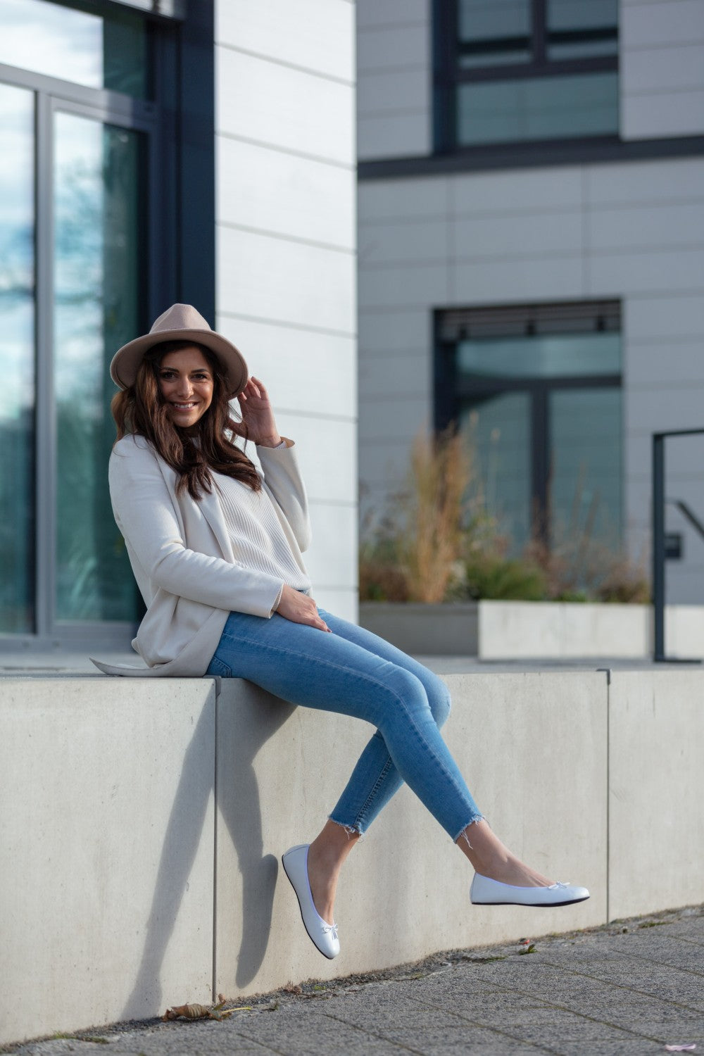 Eine Frau in weißem Blazer, LOOQ White Top von ZAQQ, Jeans und beigem Hut sitzt auf einem Betonvorsprung vor einem modernen Gebäude, lächelt und hält mit einer Hand ihren Hut. Sie sieht entspannt und lässig aus, mit übereinandergeschlagenen Beinen und Ballerinas aus Nappaleder an den Füßen.