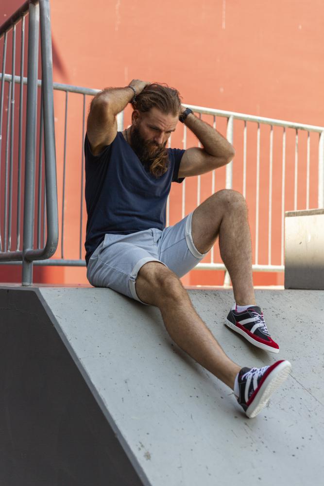 Ein Mann mit Bart sitzt auf einem Betonhang und hat die Hände auf dem Kopf. Er trägt ein marineblaues Hemd, hellgraue Shorts und leuchtend rote Turnschuhe aus der OAQLEY-Kollektion von ZAQQ. Im Hintergrund ragt eine rote Wand mit einem Metallgeländer hervor.