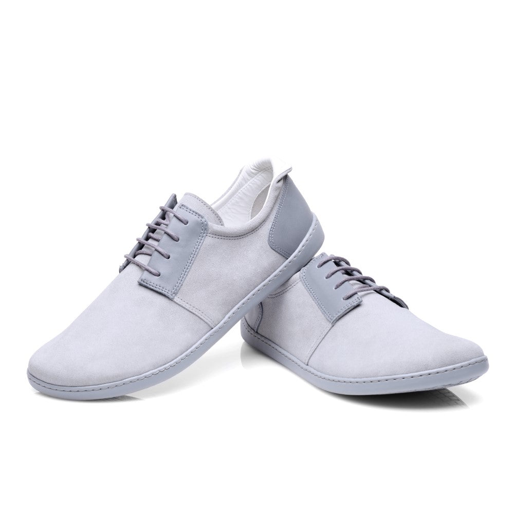 Wir präsentieren die PIQUANT Lightgrey Sneaker von ZAQQ: Diese lässigen Sneaker präsentieren sich in minimalistischem Design aus hochwertigem Veloursleder. Ein Schuh steht aufrecht, der andere liegt auf der Seite und hebt einen etwas dunkleren grauen Akzent an Ferse und Zunge hervor. Die passende graue Sohle rundet diesen stylischen Barfußschuh-Look perfekt ab.