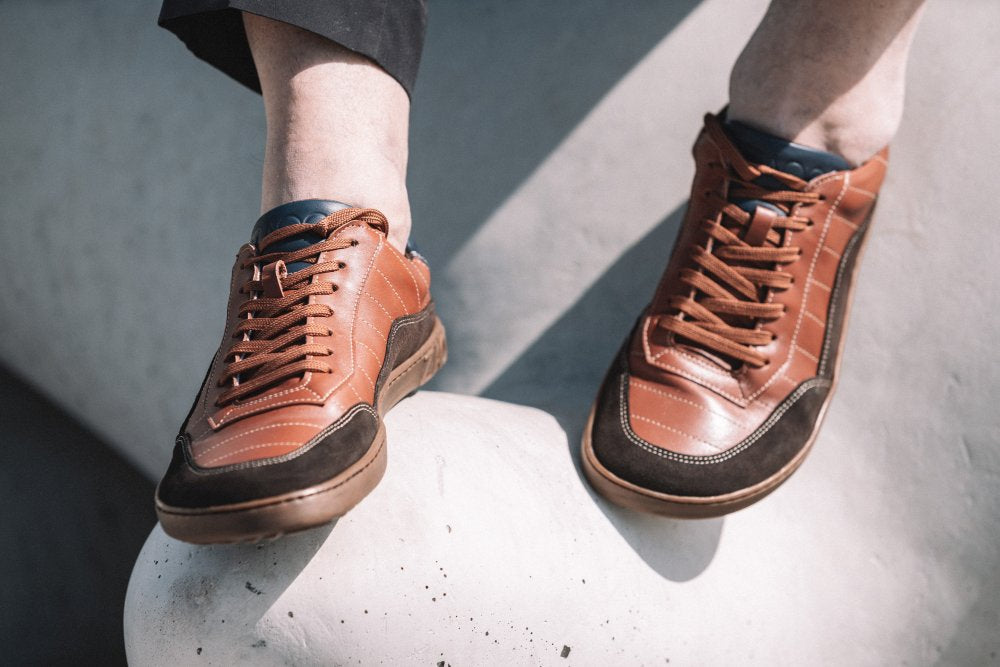 Nahaufnahme einer Person, die den QAMPION Brown Sneaker von ZAQQ mit dunkelbraunen Akzenten und flexibler Gummisohle trägt und auf einer weißen Oberfläche sitzt. Die Beleuchtung wirft weiche Schatten und hebt die Schuhdetails hervor.