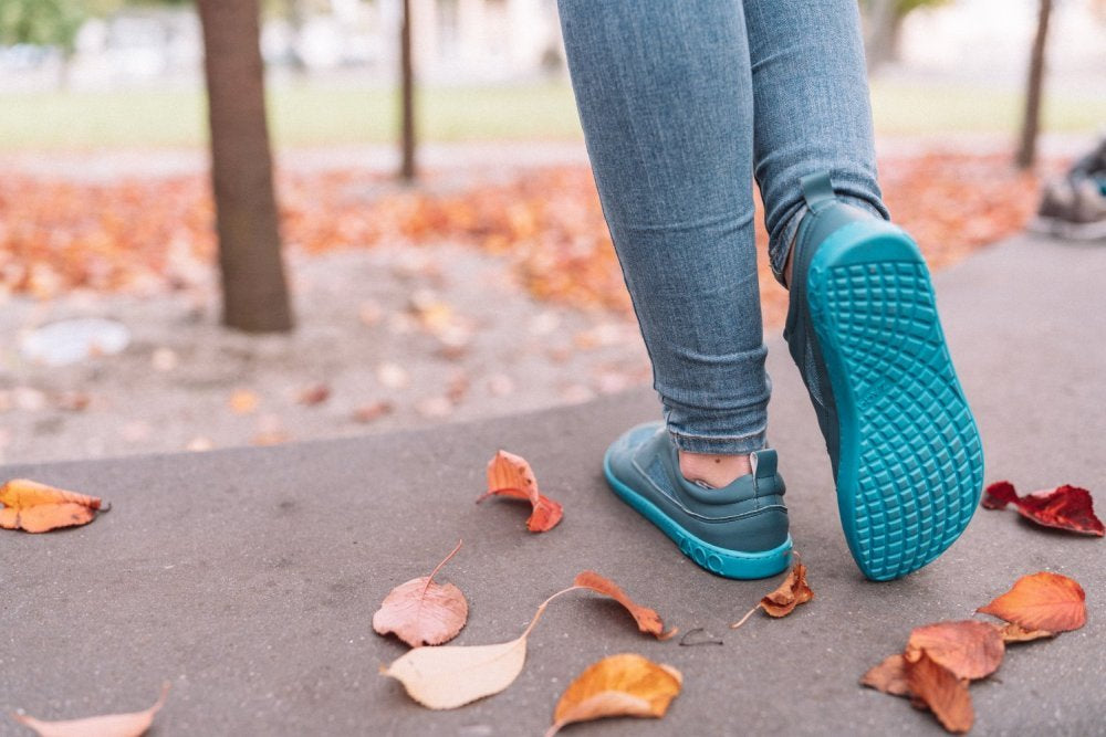 Eine Person, die ZAQQs QARLY Turquoise Sneakers und Jeans trägt, schlendert einen mit leuchtenden Herbstblättern übersäten Weg entlang. Bäume säumen den Weg und das sanfte Licht lässt auf einen ruhigen, bewölkten Tag schließen.