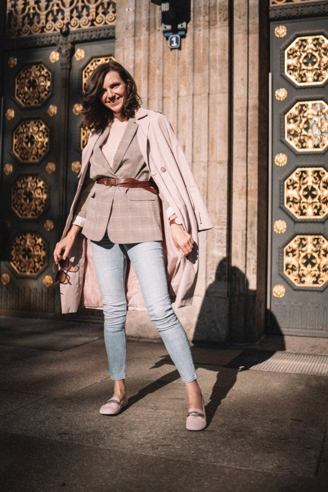 Eine stilvolle Frau posiert selbstbewusst auf einer sonnenbeschienenen Straße. Sie trägt einen hellkarierten Blazer mit Gürtel in der Taille, dazu hellblaue Jeans und rosafarbene ZAQQ QERP Candy-Loafer aus Veloursleder. Ihr Trenchcoat liegt über ihren Schultern, vor dem Hintergrund verzierter Türen.