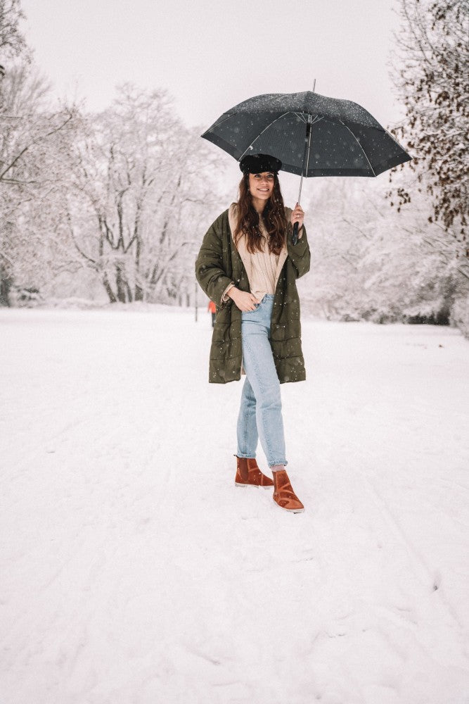 Eine Person in einem grünen Mantel und blauen Jeans steht auf einem verschneiten Weg und hält einen schwarzen Regenschirm. Sie trägt Schuhe in ZAQQ QEY WEST Cognac und einen schwarzen Hut. Schneebedeckte Bäume bilden die perfekte Winterkulisse.