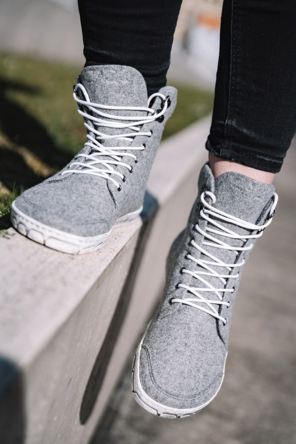 Nahaufnahme der QICE Grey High-Top-Sneakers von ZAQQ mit weißen Schnürsenkeln und Outdoor-Sohle, getragen von einer Person, die auf einem Betonvorsprung balanciert. Die Schuhe sind aus einem weichen, strukturierten Material gefertigt und die Person trägt eine schwarze Hose.