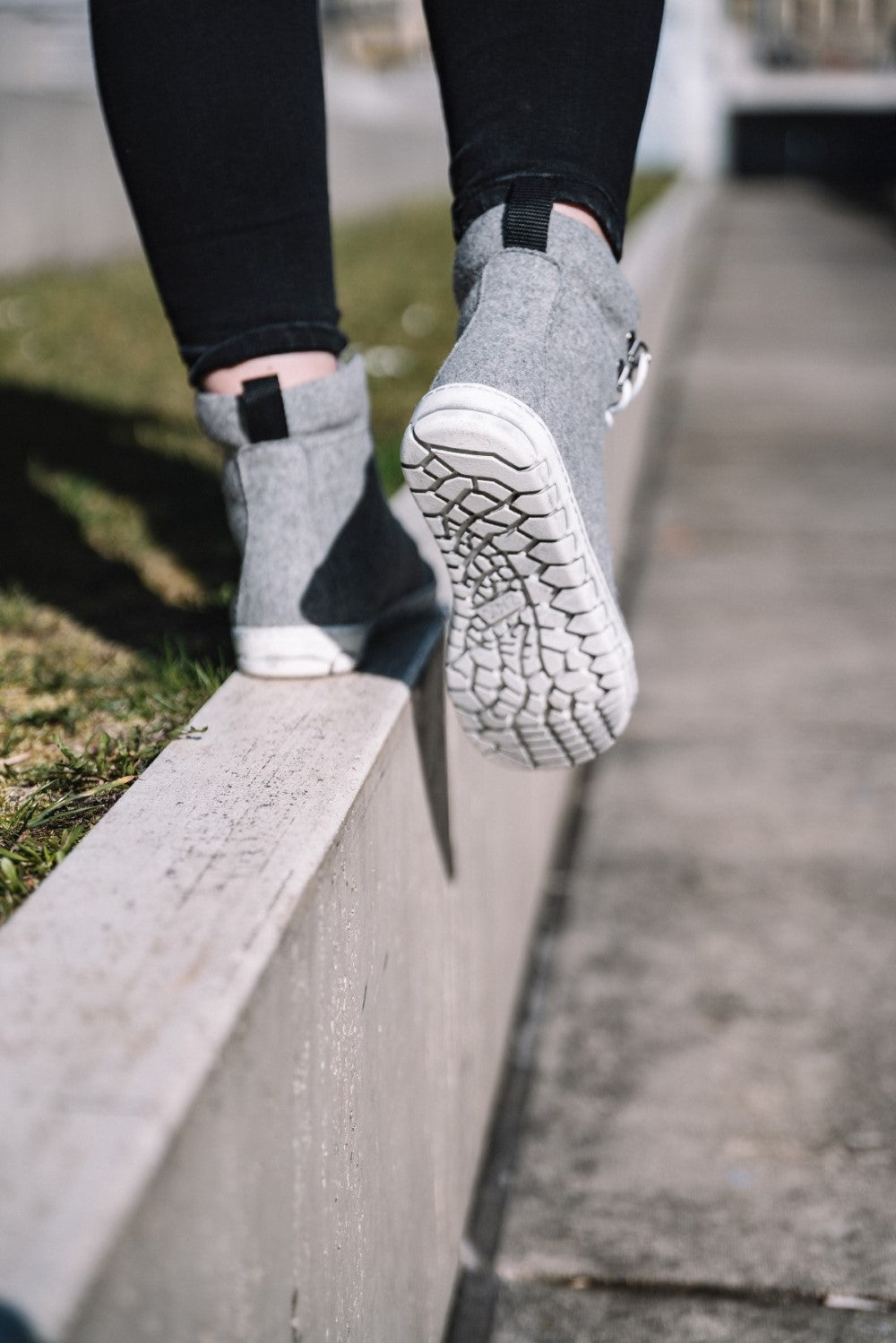 Eine Person balanciert auf einem schmalen Felsvorsprung und trägt die QICE Grey High-Top-Sneaker mit weißer Sohle von ZAQQ. Sie zeigen die perfekte Mischung aus Stil und Komfort, die an Barfußschuhe erinnert. Im Hintergrund ist eine verschwommene Ansicht eines Gehwegs und Grasses zu sehen.