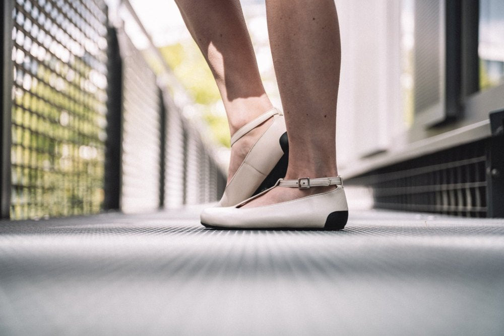 Nahaufnahme einer Person, die auf Zehenspitzen in den ZAQQ QITE Beige Ballerinas steht, gefertigt aus hochwertigem Nappaleder. Die Schuhe verfügen über zarte Knöchelriemen, die den verschwommenen Hintergrund aus Metallgeländern und üppigem Grün wunderschön betonen.