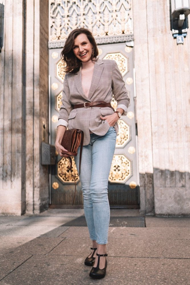 Eine Frau in kariertem Blazer und hellen Jeans steht selbstbewusst neben einer verzierten Haustür, den Gürtel in der Taille festgeschnallt. Mit einer QITE Dark Brown-Handtasche von ZAQQ über der Schulter und kurzen braunen Haaren, in denen die Sonne warm schimmert, strahlt sie Eleganz aus, wie eine selbstbewusste Ballerina, die bereit ist, die Welt zu umarmen.