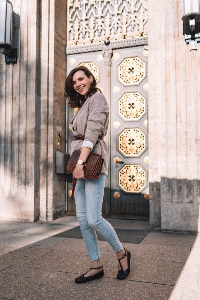 Eine Frau steht vor einer verzierten Tür mit geometrischen Mustern, gekleidet in einen grauen Blazer, blaue Jeans und schwarze Schuhe aus Glattleder. Sie trägt die QITE Dark Brown Ledertasche von ZAQQ, während die sonnenbeschienene Szene Schatten auf den Boden wirft.