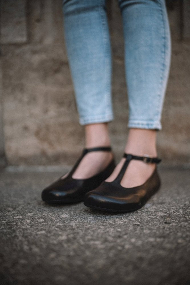 Eine Person in hellblauen Jeans und ZAQQs dunkelbraunen Schuhen QITE mit Barfußsohle steht auf einer strukturierten Betonoberfläche. Der Hintergrund ist eine verschwommene, neutral getönte Wand.