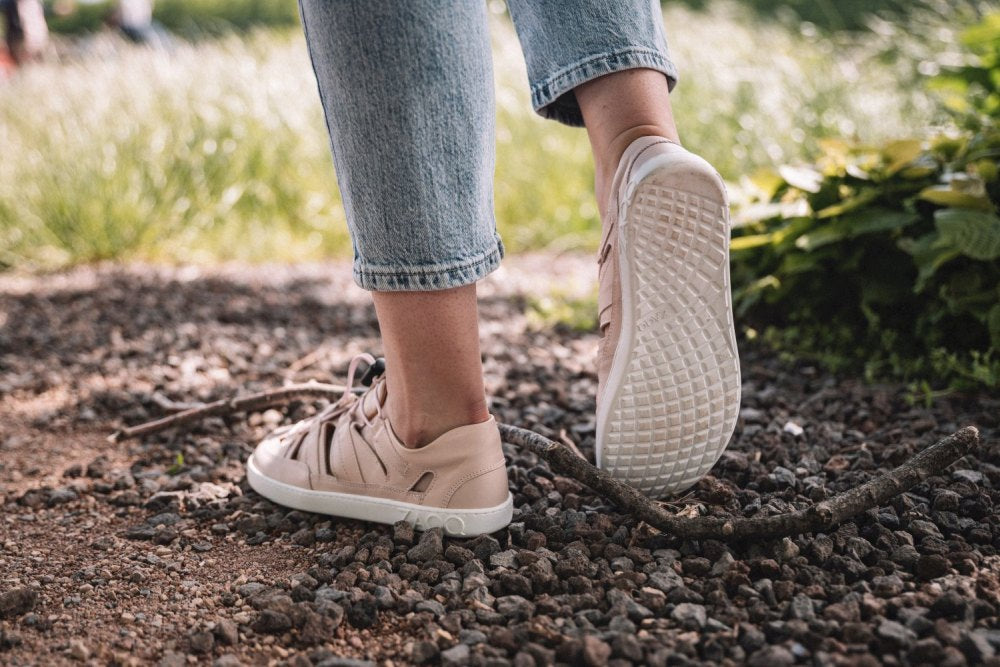 Eine Person in hellrosa QLEAR Sneakers von ZAQQ und Blue Jeans läuft auf einem Schotterweg. Der Boden, der hervorragenden Halt bietet, ist mit kleinen Steinen und einem gebogenen Stock übersät, im Hintergrund ist grünes Gras zu erkennen.