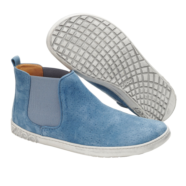 Die QOLLINS Blue Slipper von ZAQQ sind aus blauem Veloursleder gefertigt und haben weiße Gummisohlen mit strukturiertem Griffmuster. Sie sind auf Komfort ausgelegt und verfügen über elastische Seitenteile für einfaches Tragen sowie ein dezentes Logo in der Nähe der Ferse. Ein Schuh wird aufrecht präsentiert, während der andere seine Sohle zur Schau stellt.