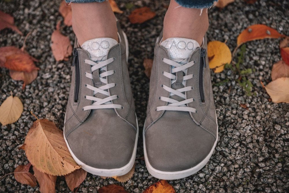 Eine Person, die die ZAQQ QOMFY Wide Grey Sneaker mit weißer Sohle trägt, steht auf einem Kiesweg, umgeben von Herbstlaub. Die Schuhe haben weiße Schnürsenkel und seitliche Reißverschlüsse und vereinen Stil mit Komfort. Die Knöchel sind über den Sneakern sichtbar, wobei ausgefranste Jeanskanten für einen lässigen Touch sorgen.