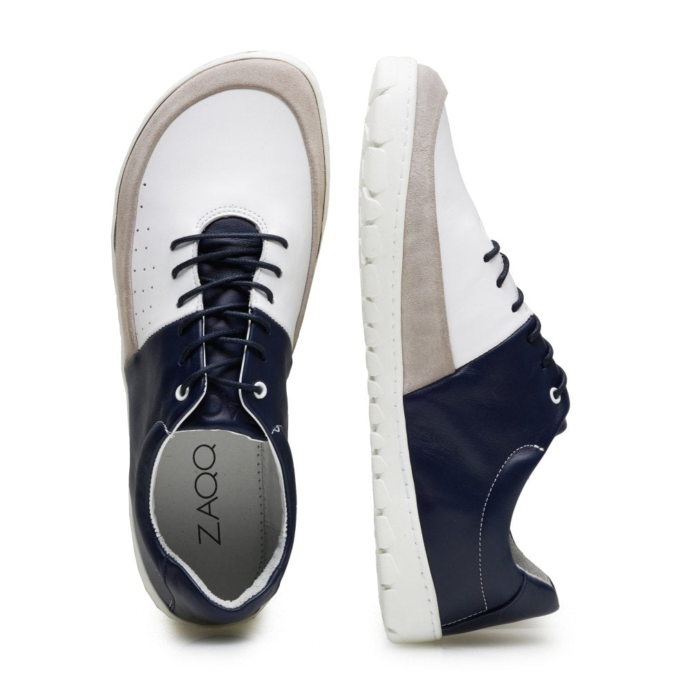 Wir präsentieren den QOOL Navy von ZAQQ, ein Paar stylischer Barfuß-Sneaker mit modernem Design. Ein Schuh steht aufrecht und präsentiert sein marineblaues und weißes Nappaleder mit grauen Akzenten, während der andere horizontal liegt, um die Sohle und den ZAQQ-Markennamen auf der Innensohle hervorzuheben. Erleben Sie unvergleichlichen Tragekomfort bei jedem Schritt.