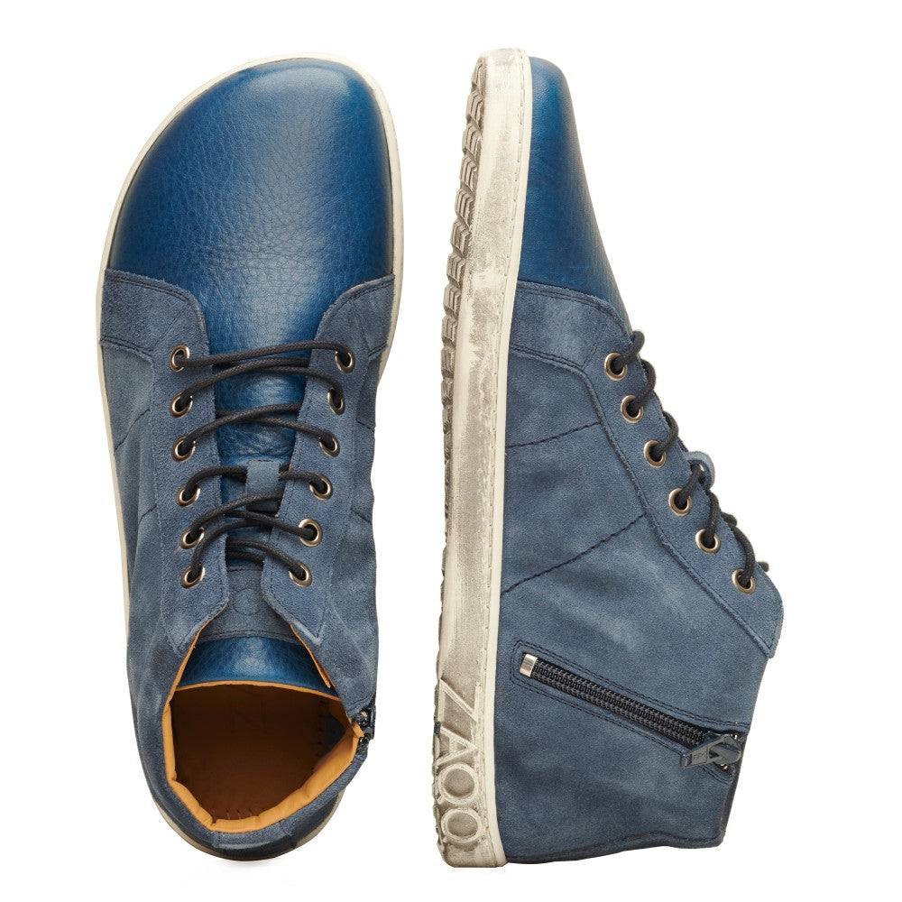 Zwei ZAQQ QORE Mid Deep Sea High-Top-Schuhe aus blauem Leder, wobei ein Schuh nach oben und der andere von der Seite zeigt. Die Schuhe verfügen über schwarze Schnürsenkel, die durch silberne Ösen und Veloursleder-Einsätze verflochten sind. Ein Reißverschluss ziert die Seite eines Schuhs, während die Sohle den Markennamen ZAQQ prominent zur Schau stellt.