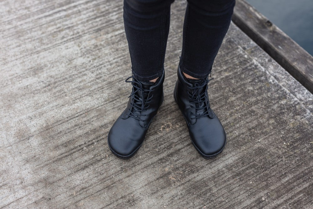 Eine Person in schwarzen Röhrenjeans und QUINTIC Winter Vegan Black-Schuhen von ZAQQ steht auf einem verwitterten Holzsteg neben dem Wasser. Der Winkel fängt die Unterschenkel und Füße ein und betont die Textur der Stiefel aus wasserabweisendem Material und der Bretter darunter.