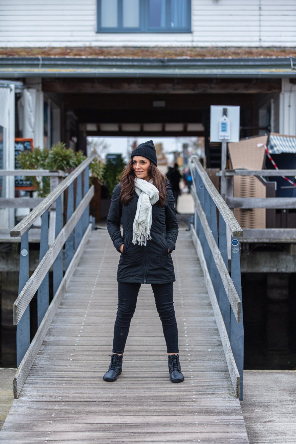 Eine Frau steht auf einem Holzsteg, die Hände in den Taschen, trägt einen schwarzen Mantel und eine schwarze Hose, ergänzt durch eine schwarze Strickmütze. Ihr weißer Schal ist anmutig drapiert, während sie die QUINTIC Winter Vegan Black Schuhe von ZAQQ präsentiert, die aus wasserabweisendem Material hergestellt sind. Im Hintergrund ist hinter den mit Pflanzen gesäumten Geländer ein Gebäude zu sehen.