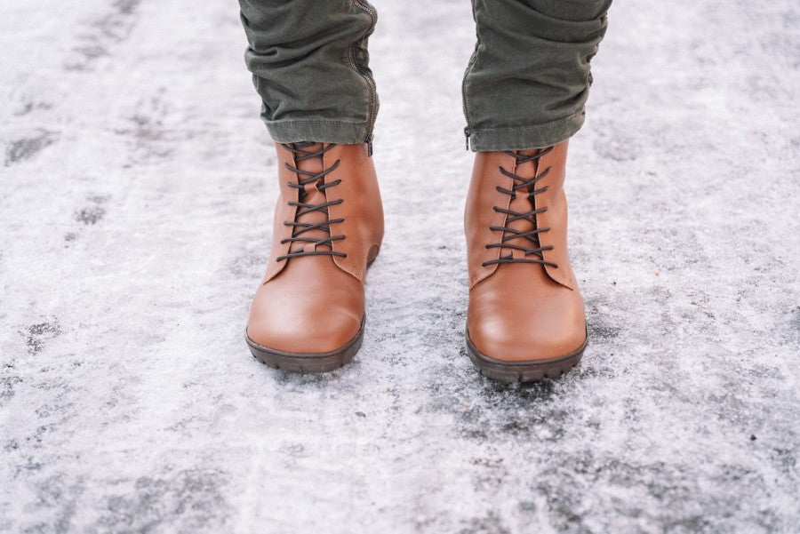 Eine Nahaufnahme zeigt eine Person, die ZAQQs QUINTIC Winter Vegan Tabaco Stiefel mit dunklen Kautschuksolen und dunkelgrünen Hosen trägt und auf einer schneebedeckten Fläche steht. Die ordentlich gebundenen Schnürsenkel und der verdichtete Schnee, durchsetzt mit Eisflecken, evozieren eine winterliche Szene.