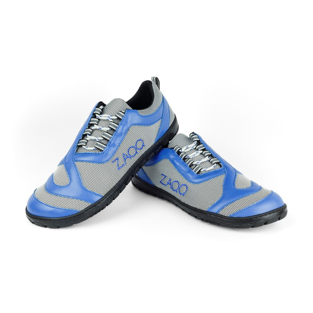 Ein Paar ZAQQ QUIQ Trail Blue Sportschuhe präsentiert ein sportliches Barfußschuh-Design mit atmungsaktivem Meshgewebe-Material und schwarzer Sohle. Der Markenname ZAQQ prangt seitlich, da die Schuhe kunstvoll auf einem weißen Hintergrund dargestellt sind und aneinander gelehnt sind.