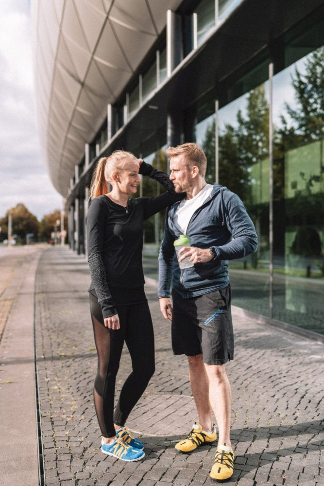 Ein Mann und eine Frau in Sportkleidung stehen vor einem modernen Gebäude mit großen Glasfenstern. Der Mann hält eine Tasse und sie scheinen in ein Gespräch vertieft zu sein. Beide tragen die RIQ Yellow Laufschuhe von ZAQQ, die für ihr flexibles Tragegefühl auf dem gepflasterten Gehweg bekannt sind.