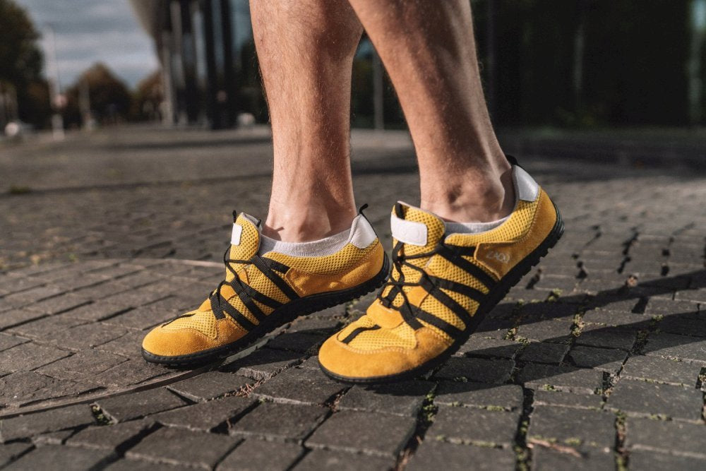 Eine Person, die RIQ Yellow-Schuhe mit schwarzen Schnürsenkeln von ZAQQ trägt, schlendert auf einem Kopfsteinpflasterweg entlang. Diese Barfußschuhe zeichnen sich durch ein minimalistisches, leichtes Design aus und bieten ein flexibles Tragegefühl. Die Sonne wirft lange Schatten und lässt vermuten, dass es entweder früher Morgen oder später Nachmittag ist.