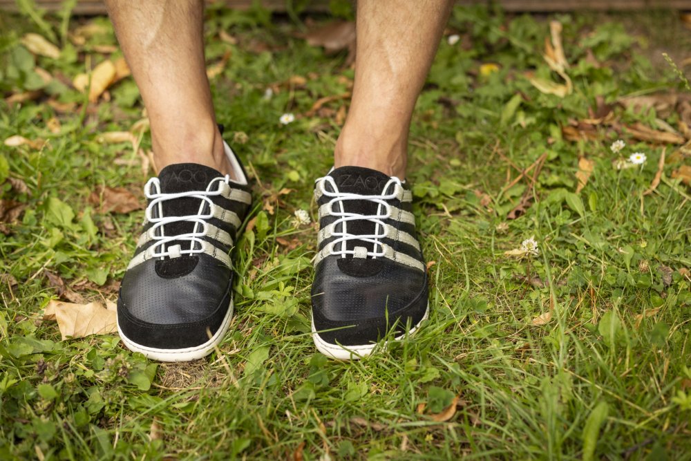 Eine Nahaufnahme zeigt jemanden, der die SPARQ Low Black White Sneaker von ZAQQ trägt. Diese Schuhe aus weichem Nappaleder weisen markante weiße Schnürsenkel und Streifen auf. Die Person steht auf Gras, das mit verstreuten Blättern und kleinen weißen Blumen übersät ist.