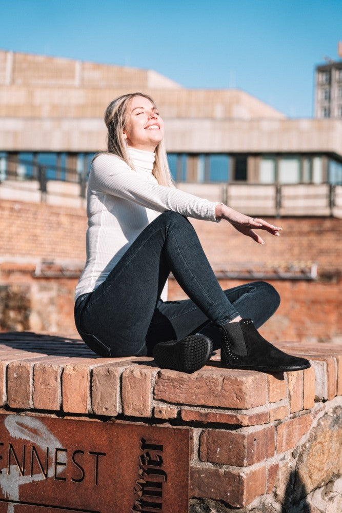 Eine Frau mit langen blonden Haaren sitzt auf einer Backsteinmauer unter einem klaren blauen Himmel und lächelt mit geschlossenen Augen. In einen weißen Rollkragenpullover, dunkle Jeans und die SPARQLE Shine Velours Black Ankle Boots von ZAQQ gekleidet, genießt sie mühelos das Sonnenlicht in einem Moment minimalistischer urbaner Gelassenheit.