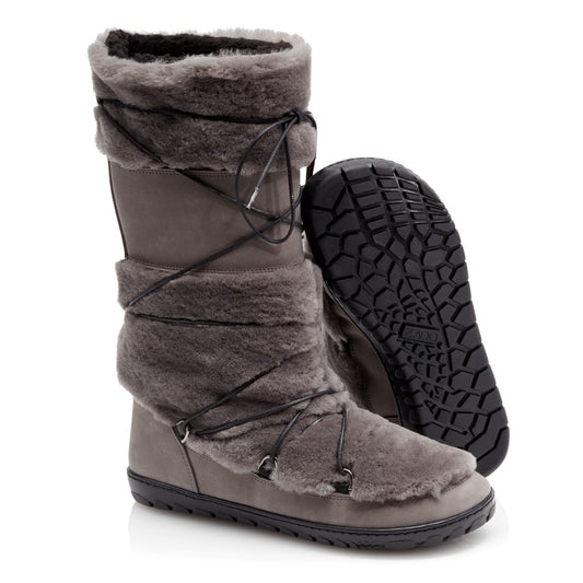 Ein Paar TORQ-Winterstiefel von ZAQQ in grauem, pelzgefüttertem Design mit schwarzen Schnürsenkeln. Ein Stiefel steht aufrecht, während der andere leicht geneigt ist, um die strukturierte Sohle zu präsentieren und ein Barfußgefühl zu vermitteln. Die Außenseite besteht aus glattem Material mit Pelzbesatz.