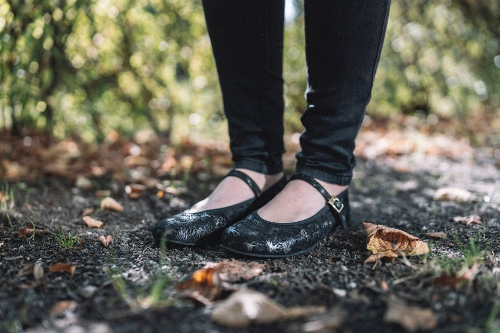 Eine Nahaufnahme einer Person in eleganten schwarzen TWIST Ennis-Schuhen von ZAQQ, kombiniert mit dunklen Jeans, die auf einem Kiesweg steht. Gefallene Blätter und Grün bereichern die ruhige, natürliche Umgebung und erinnern an die Anmut einer Ballerina inmitten der Natur.