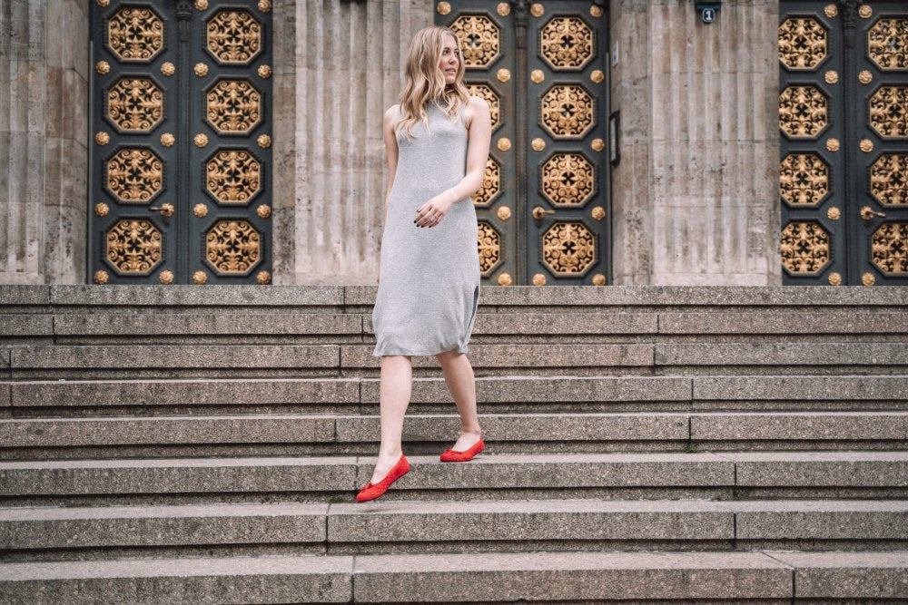 Eine Frau mit langen blonden Haaren, einem ärmellosen grauen Kleid und auffälligen roten ZAQQ TWIST-Schuhen, geht anmutig Steintreppen hinunter. Im Hintergrund sind verzierte Türen mit goldenen Akzenten und hohen Säulen zu sehen, die der Szene einen Hauch von Eleganz verleihen.