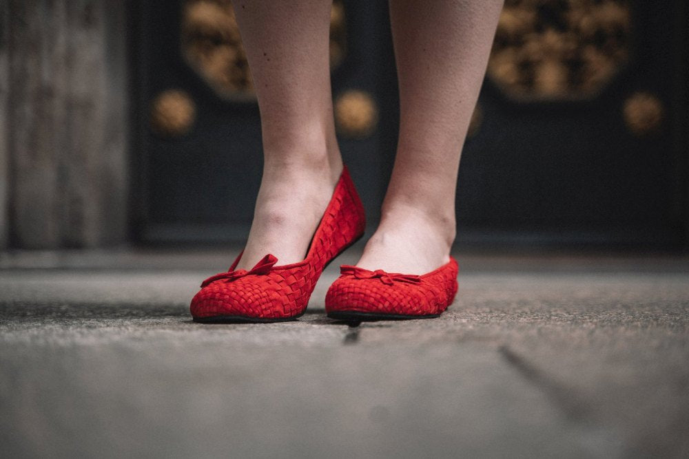 Nahaufnahme der Beine einer Person, die die TWIST Red Ballerinas von ZAQQ trägt, mit strukturiertem Design und Schleifen an der Oberseite. Die Schuhe aus Velourleder stehen auf grauem Beton, vor einem dunklen Hintergrund, der mit kunstvollen goldenen Details verziert ist.