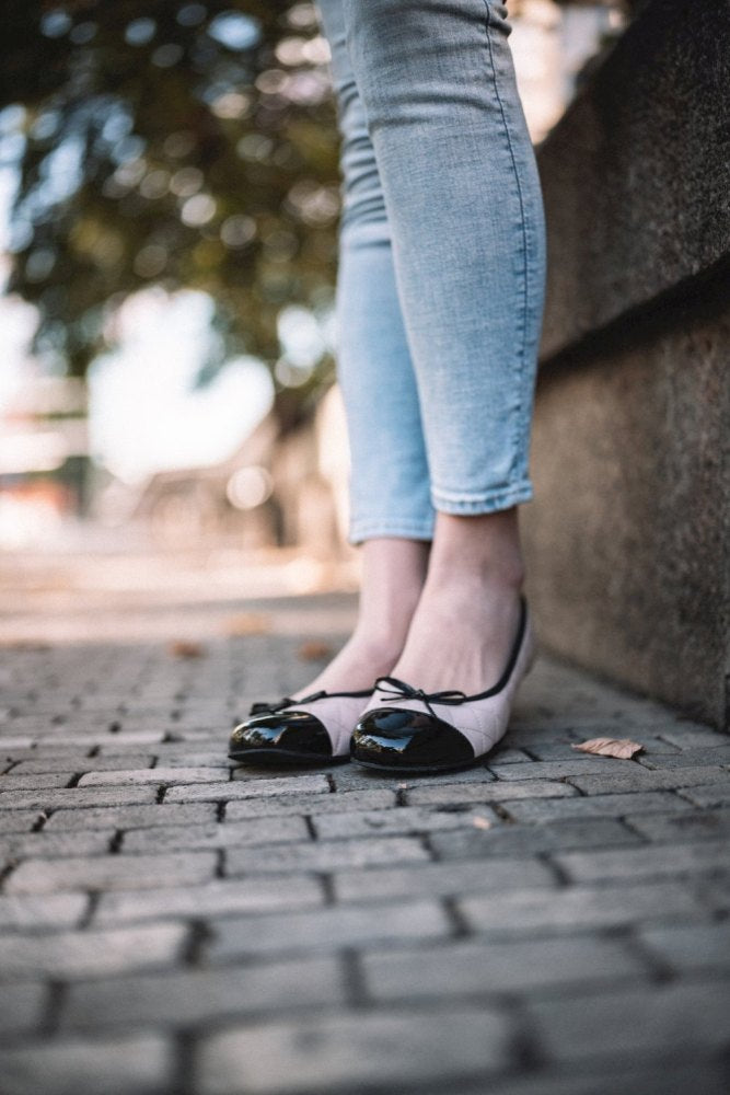 Nahaufnahme einer Person auf einem Kopfsteinpflasterweg, die hellblaue Jeans und schwarze TWIST Varnish Nude-Ballerinas von ZAQQ trägt, die mit Schleifen verziert sind. Der Weg ist von einer Steinmauer gesäumt und verschwommenes Grün verleiht dem Hintergrund Charme.