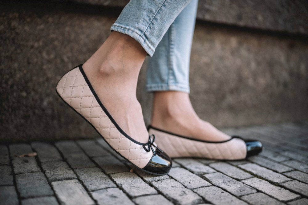 Eine Person trägt hellblaue Jeans und ZAQQs TWIST Varnish Nude-Schuhe, beige gesteppte Ballerinas mit schwarzer Lackkappe, und steht auf einem Kopfsteinpflaster. Die Schuhe sind aus Kautschuksohlen gefertigt und betonen einen lässigen und doch stilvollen Look, der auf Komfort und Haltbarkeit ausgerichtet ist.